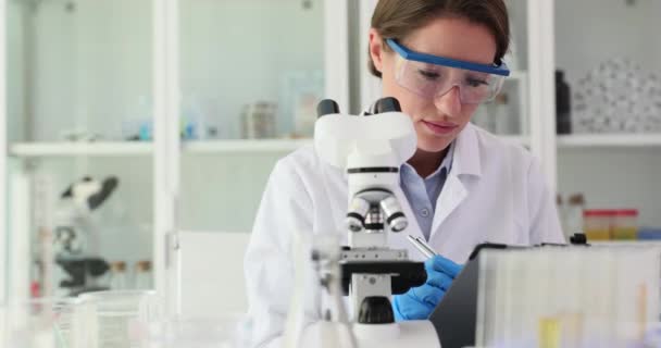 El científico mira a través de un microscopio y escribe información sobre un experimento o investigación en laboratorio. Experimentos químicos - Imágenes, Vídeo