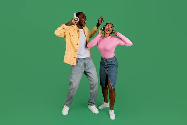 Funny young african american couple wearing casual outfits listening to music and dancing, happy black man and woman using modern wireless headphones, enjoying favorite songs, green background - Photo, Image