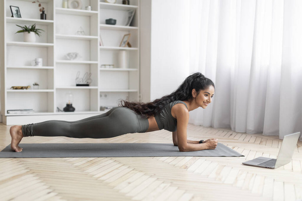 Fegyelmezett és odaadó, egy fiatal hindu nő edzőruhában elmerül egy energikus gyakorlat rutin, hölgy planking által vezetett fitness oktató keresztül laptopját otthon, oldalnézet - Fotó, kép