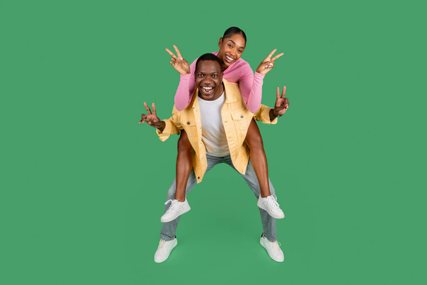 Nettes glückliches schönes afrikanisches Millennial-amerikanisches Paar hat Spaß auf grünem Studiohintergrund. Positive hübsche junge schwarze Frau huckepack ihren Freund, zeigt Friedensgeste und lächelt - Foto, Bild