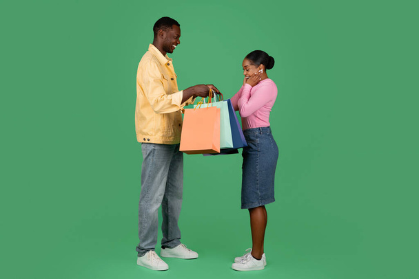 Cool bello alto millenario ragazzo afroamericano rende felice la sua bella fidanzata emotiva, dando i suoi acquisti borse di carta colorate, isolato su sfondo studio verde, lunghezza intera - Foto, immagini