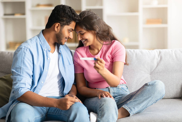 Junges indisches Paar strahlt Freude aus, während es sich auf dem Sofa bequem macht, zärtliche Glücksmomente teilt, einen positiven Schwangerschaftstest hält und ihre Stirn berührt - Foto, Bild