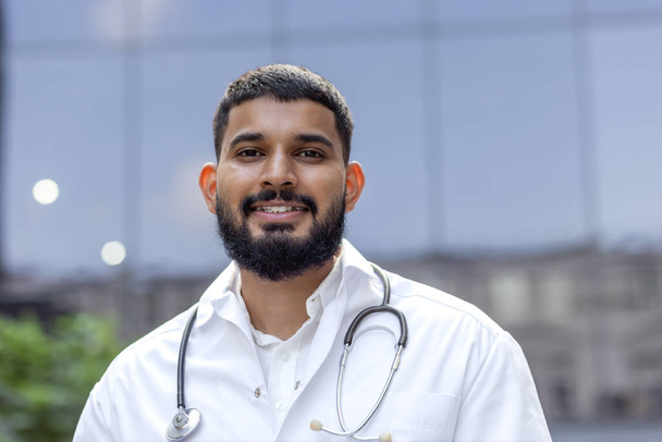 Portrait en gros plan d'un jeune médecin musulman, étudiant debout dehors en manteau blanc et tenant un stéthoscope, regardant avec confiance et souriant la caméra. - Photo, image