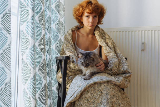 Girl hugging cat, sitting in blanket home, frozen. teen barefoot, red-haired, curly, morning, wrapped in blanket, sits hugging domestic cat, near heating radiator, large window covered with curtains. - Photo, Image