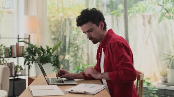 Vue latérale du jeune entrepreneur en vêtements décontractés travaillant sur le rapport tout en étant assis à la maison avec un ordinateur portable - Séquence, vidéo