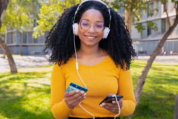 Kulaklık takan mutlu siyah kadın akıllı telefondan kredi kartıyla ödeme yapıyor. Kameraya bakıyorum. Konsept: çevrimiçi mağaza ödemesi - Fotoğraf, Görsel