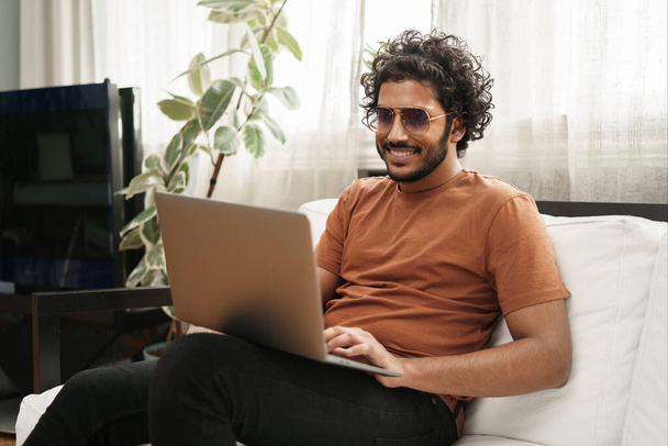 Gelukkig mannelijk student werken op een laptop terwijl gezeten op een bank in een gezellige kamer - Foto, afbeelding