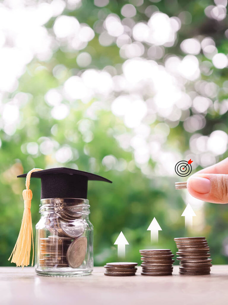 Close up hand putting coin in stack of coins with target icon and arrow up icons, Glass bottle with graduation hat. The concept of saving money for education, student loan, scholarship, tuition fees in New Year 2024 - Photo, Image