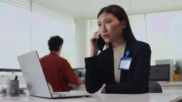 Közepes felvétel női irodai dolgozó beszél okostelefonon, miközben ül az asztalnál vezeték nélküli laptop - Felvétel, videó