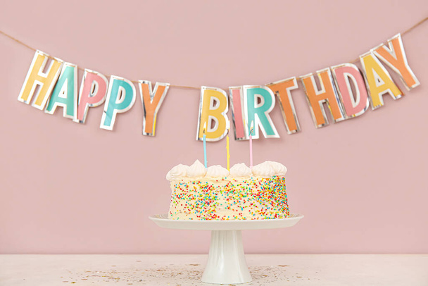 Guirnalda de fiesta y de pie con delicioso pastel de cumpleaños en la mesa cerca de la pared rosa - Foto, imagen