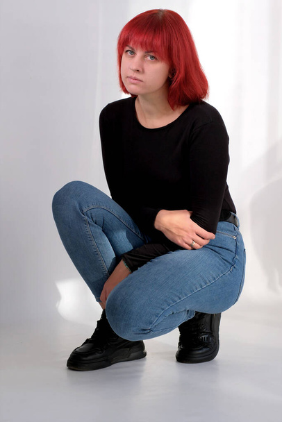 Una giovane donna attraente con una breve acconciatura arancione in una t-shirt nera e jeans in posa in studio su uno sfondo bianco. - Foto, immagini