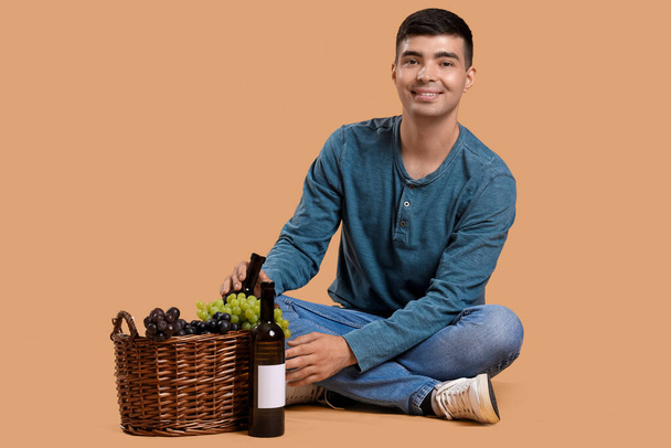 Νεαρός άνδρας με κρασί και καλάθι κάθεται σε μπεζ φόντο - Φωτογραφία, εικόνα