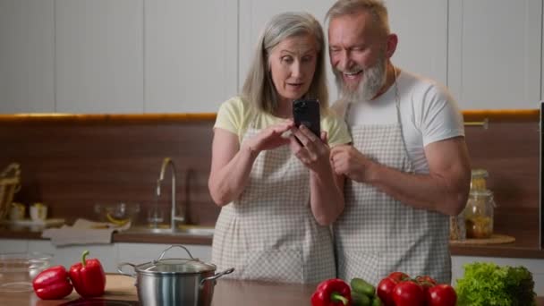 Caucasian old family mature love couple smiling middle-aged man woman use phone order healthy food with mobile delivery online looking at recipe on smartphone easy cooking prepare breakfast in kitchen - Footage, Video