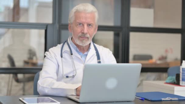 Médico mayor sonriendo en la cámara mientras trabaja en el ordenador portátil - Metraje, vídeo