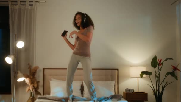 Despreocupado hilarante chica afroamericana mujer étnica saltando en la cama en casa divirtiéndose celebrando vacaciones divertida noche de baile fiesta en casa cantando canción en el teléfono móvil escuchando música en auriculares - Imágenes, Vídeo