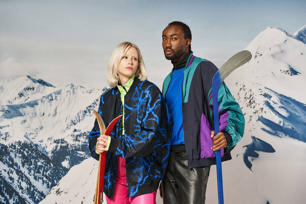 stylish multicultural couple in vibrant clothes posing with skis and hockey stick, winter concept - Fotografie, Obrázek