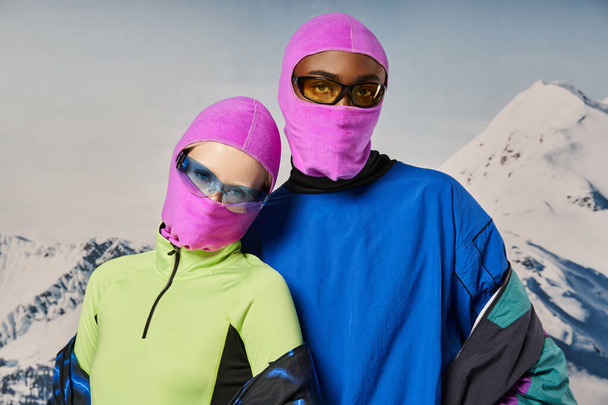 young stylish couple in warm vibrant outfits and pink balaclavas with snowy backdrop, winter concept - Photo, Image