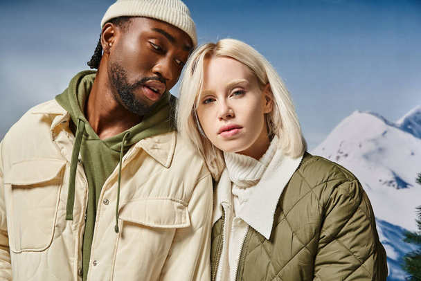 attractive couple in stylish modern outfits with snowy mountain on backdrop, winter concept - Zdjęcie, obraz