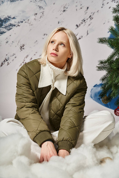 красивая блондинка в теплой зимней куртке сидит на снегу и мечтательно глядя в сторону, мода - Фото, изображение