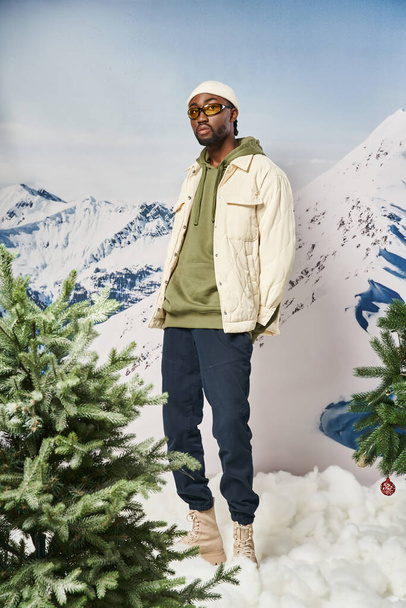 fashionable man in stylish warm outfit with beanie hat posing next to pine trees, winter concept - Foto, afbeelding
