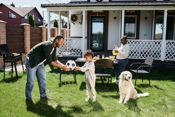 οικογενειακό Σαββατοκύριακο, ευτυχισμένος Αφροαμερικάνος πατέρας δίνοντας μπάλα ποδοσφαίρου στο γιο κοντά στη γυναίκα που στρώνει το τραπέζι - Φωτογραφία, εικόνα
