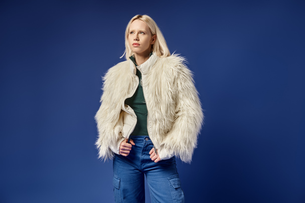 dreamy and stylish blonde woman in faux fur jacket and denim jeans posing on blue backdrop - Фото, изображение