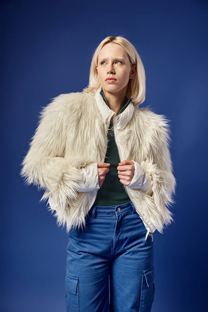 pensive and stylish blonde woman in faux fur jacket and denim jeans posing on blue backdrop - 写真・画像