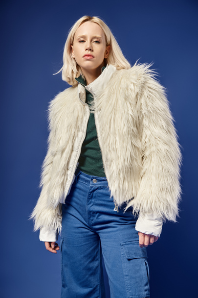 winter fashion, attractive blonde woman in faux fur jacket and denim jeans posing on blue backdrop - 写真・画像