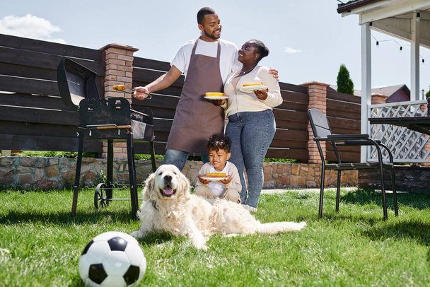 joyful african american family holding plates with grilled corn near bbq grill and dog on backyard - Foto, Bild