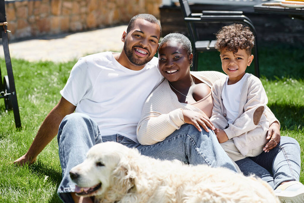 Familienporträt einer positiven afrikanisch-amerikanischen Familie, die in die Kamera blickt und neben Hund auf dem Rasen sitzt - Foto, Bild