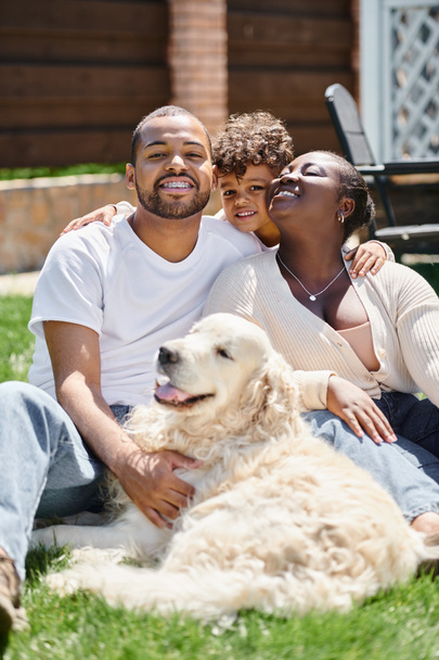 familie portret van vrolijke Afrikaanse Amerikaanse ouders en zoon glimlachend en zittend op gazon in de buurt van hond - Foto, afbeelding