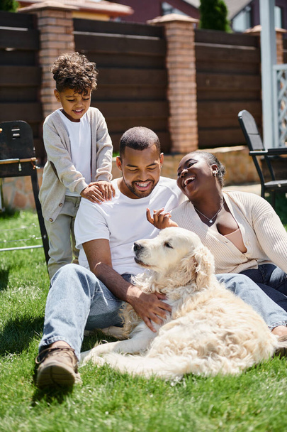 portrait of cheerful african american parents and son smiling and sitting on green lawn near dog - Photo, Image