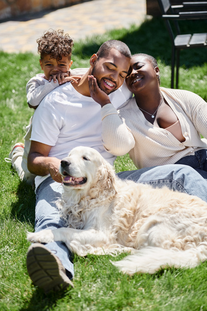 portrait of happy african american parents and son smiling and sitting on green lawn near dog - Foto, Imagen