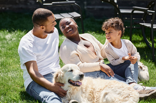 ジョリー・アフリカ系アメリカ人の両親と息子が笑顔で犬の近くの緑の芝生に座る家族の瞬間 - 写真・画像