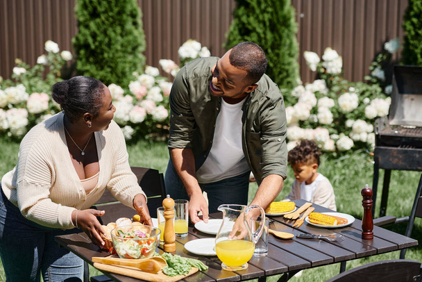 alegre afroamericano padres sirviendo mesa en jardín cerca hijo jugando en patio trasero, familia - Foto, imagen