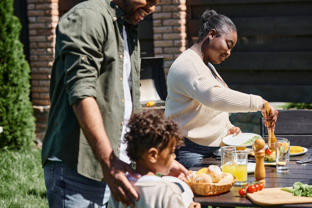 veselí afroameričtí rodiče servírující jídlo na stole v zahradě poblíž syna držícího fotbalový míč - Fotografie, Obrázek