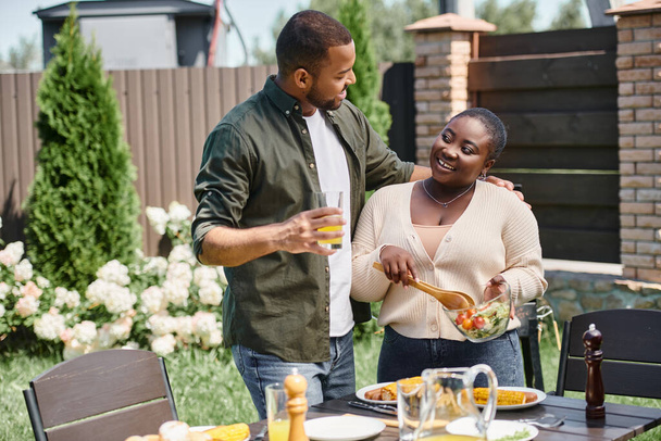 african american man hugging happy wife mixing salad in glass bowl while having bbq on backyard - Photo, Image