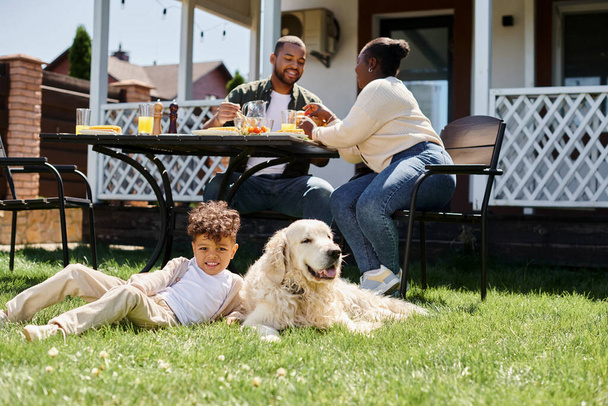 glücklich afrikanisch-amerikanisches Kind sitzt auf grünem Rasen neben Hund und Eltern beim Mittagessen im Garten - Foto, Bild