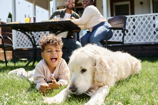 amazed african american kid sitting on green lawn near dog and parents having lunch in garden - Foto, afbeelding
