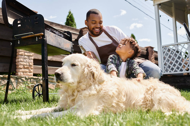 Χαρούμενος Αφροαμερικάνος με ποδιά κρατώντας λαβίδες μαγειρικής και κοιτάζοντας το σκύλο της οικογένειας κοντά στο γιο - Φωτογραφία, εικόνα