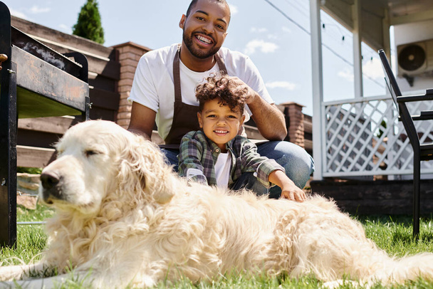 Önlüklü mutlu Afro-Amerikan adam oğlunun kıvırcık saçlarına dokunuyor ve açık havada aile köpeğinin yanında oturuyor. - Fotoğraf, Görsel