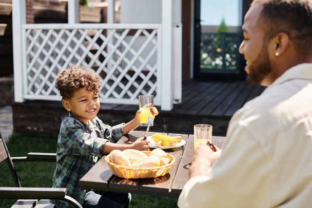 joyful african american boy eating grilled bbq meal and holding orange juice near father on backyard - Photo, Image