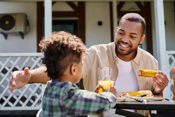 positieve Afro-Amerikaanse man met familie bbq met krullende zoon op achtertuin van huis, gegrilde maïs - Foto, afbeelding