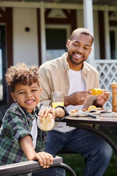 cheerful african american kid having family bbq with father on backyard of house, holding bun - Photo, Image