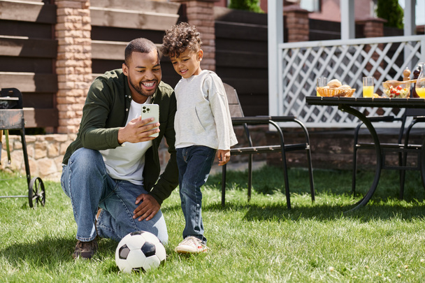 happy african american father and son looking at smartphone and standing near football on backyard - Photo, Image
