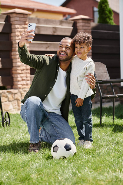 happy african american father and son looking at smartphone while taking selfie on backyard - Photo, Image