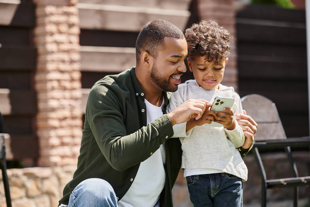 陽気なアフリカ系アメリカ人の父親と息子が家の裏庭に立ってスマートフォンを保持 - 写真・画像