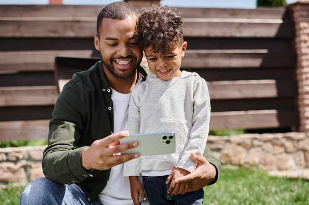 padre afroamericano satisfecho en frenos tomando selfie en teléfono inteligente con su hijo en el patio trasero - Foto, Imagen