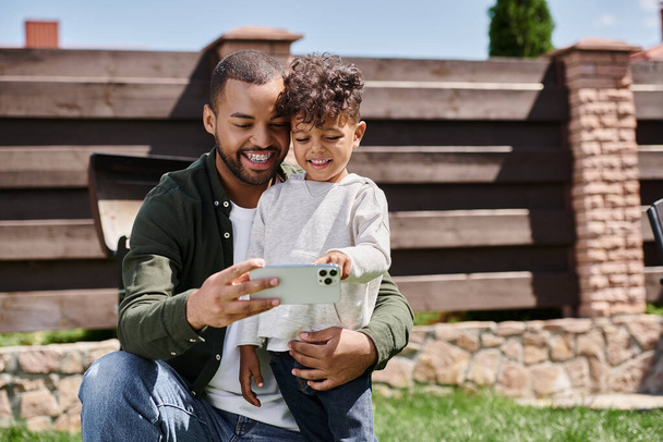 joyful african american father in braces taking selfie on smartphone with his son on backyard - Photo, Image