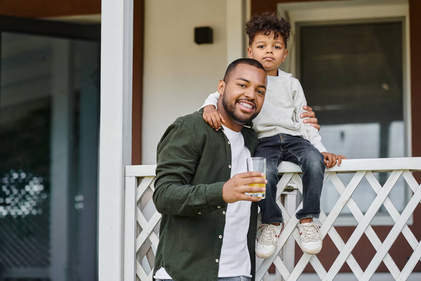 Χαρούμενος Αφροαμερικάνος πατέρας με σιδεράκια που κρατάει χυμό πορτοκάλι και αγκαλιάζει το γιο του στη βεράντα. - Φωτογραφία, εικόνα